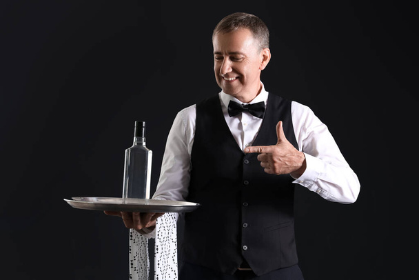 Camarero maduro apuntando a la botella de vodka sobre fondo negro - Foto, Imagen