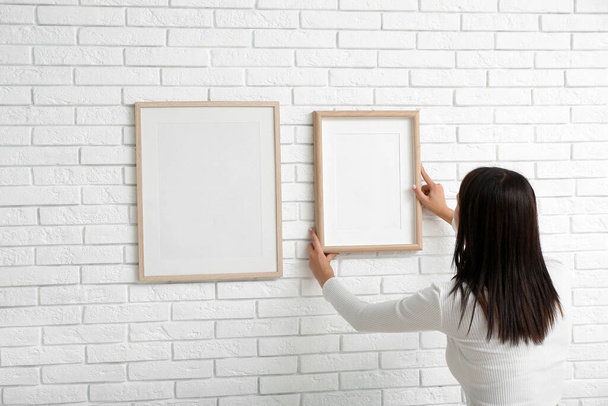 Young woman hanging blank frame on white brick wall in living room, back view - Foto, imagen