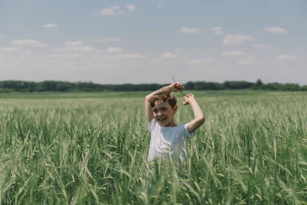 Πορτρέτο ενός χαμογελαστού αγοριού με λευκό μπλουζάκι και παίζοντας σε ένα πράσινο χωράφι με κριθάρι. Χαρούμενο παιδί που γελάει και παίζει την καλοκαιρινή μέρα. Παιδί που εξερευνά τη φύση. Καλοκαιρινή δραστηριότητα για παιδιά με ειδικές ανάγκες - Φωτογραφία, εικόνα