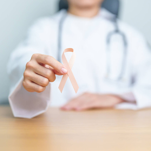 Lekarz trzymający wstążkę brzoskwiniową na miesiąc wrześniowej świadomości raka macicy. Macica i jajniki, endometrioza, histerektomia, mięśniaki macicy, rozrodczość, opieka zdrowotna i koncepcja Światowego Dnia Raka - Zdjęcie, obraz