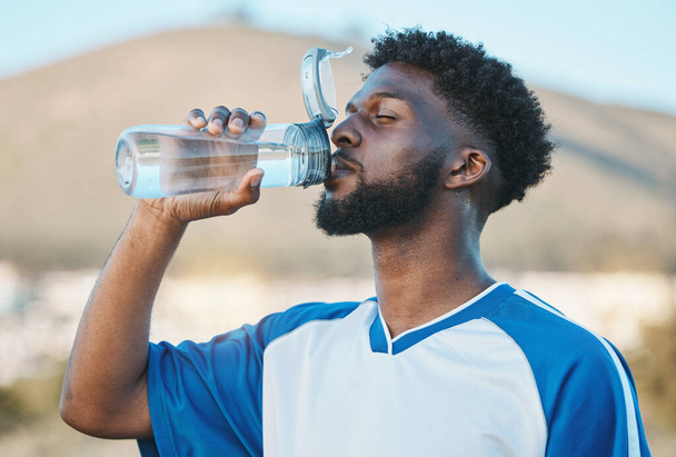 Hombre negro, fitness o jugador de fútbol bebiendo agua en el entrenamiento, ejercicio o entrenamiento en el campo de fútbol. Atleta sediento, deportivo o cansado en descanso con una bebida saludable para la energía para relajarse. - Foto, imagen