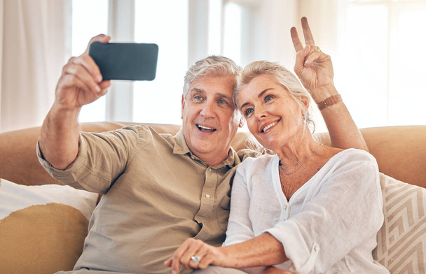 Счастливая пожилая пара, знак мира и селфи в расслаблении на диване гостиной для фотографии, памяти или влога дома. Пожилой мужчина и женщина улыбаются для фотографии, фото или социальных медиа на диване в гостиной вместе. - Фото, изображение