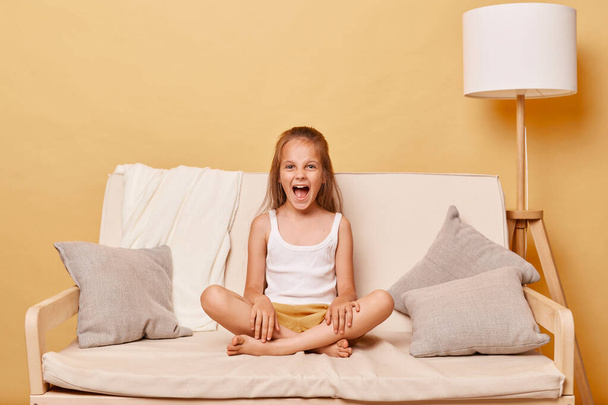Angry little girl wearing casual clothing sitting on sofa at home against beige wall having problems during teenage years having hysterical behavior screaming. - Zdjęcie, obraz