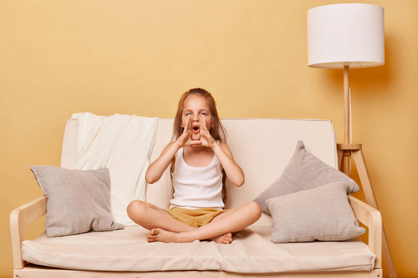 Brown haired little girl wearing casual clothing sitting on sofa at home against beige wall screaming with hands near mouth making announcement yelling loud. - Foto, Bild