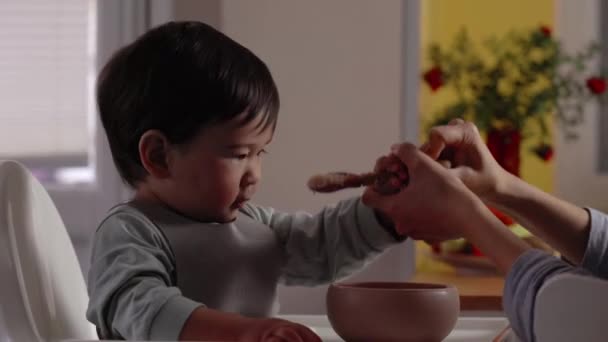 Aziatische baby probeert een lepel van zijn moeder te nemen om alleen te eten. Moeder neemt lepel en kom eten van de baby. 4k-beelden - Video