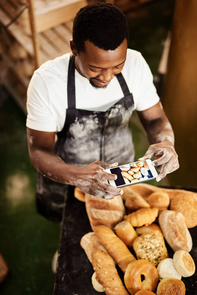 Dieses Bild ist für die heutigen Online-Backtipps. Schnappschuss eines männlichen Bäckers, der mit seinem Handy ein Foto von einer Auswahl frisch gebackener Brote macht - Foto, Bild