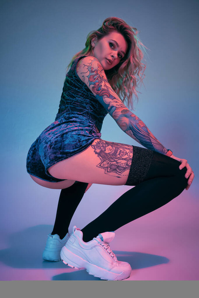 Belle jeune fille blonde tatouée en velours bleu booty shorts et t-shirt et bas noirs, faisant squats, montrant son incroyable butin - Photo, image