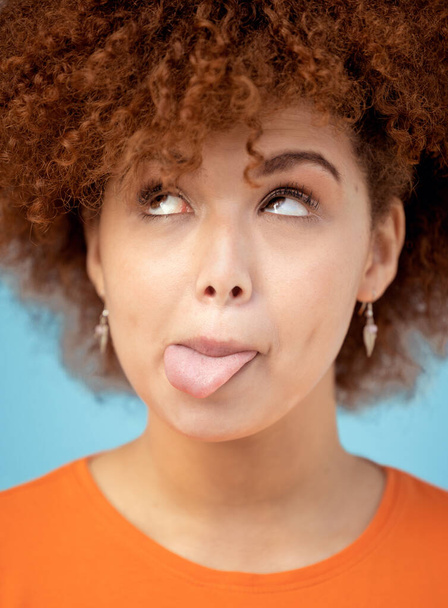 Комик, язык и лицо с чернокожей женщиной в студии на голубом фоне, которая кажется смешной или глупой. Комедия, шутка и юмор с привлекательной молодой женщиной, которая позирует в одиночестве в помещении. - Фото, изображение