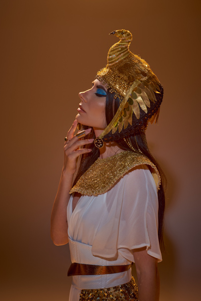 Donna in abbigliamento egiziano e copricapo toccante mento mentre posa su sfondo marrone - Foto, immagini