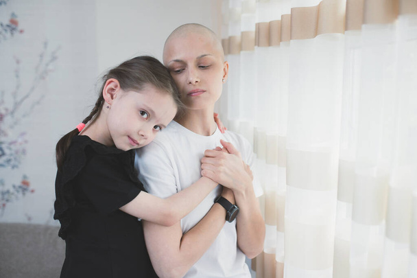 Kid girl hugs her mother with a diagnosis of leukemia near the window. Family support concept. - Foto, Imagem
