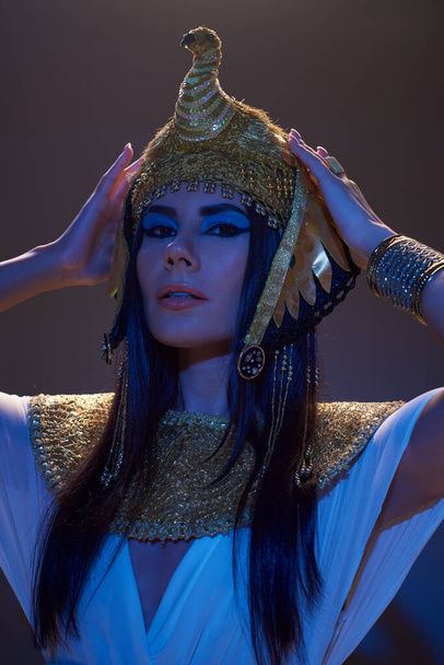 Attraente donna toccando copricapo egiziano e guardando la fotocamera in luce blu su sfondo marrone - Foto, immagini