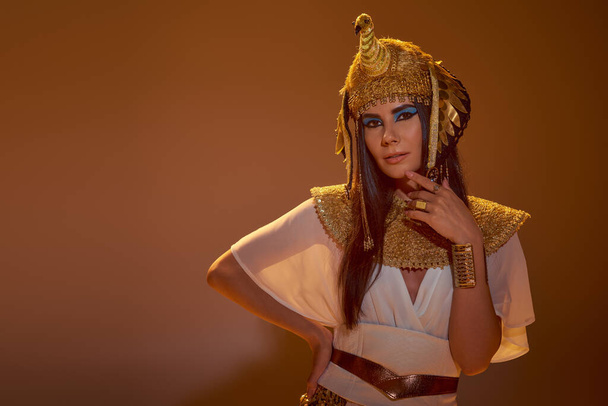Mujer con estilo en traje egipcio y tocado tocando la barbilla y posando sobre fondo marrón - Foto, Imagen