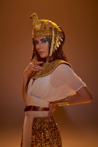 Elegante donna in look egiziano e copricapo guardando la fotocamera su sfondo marrone - Foto, immagini