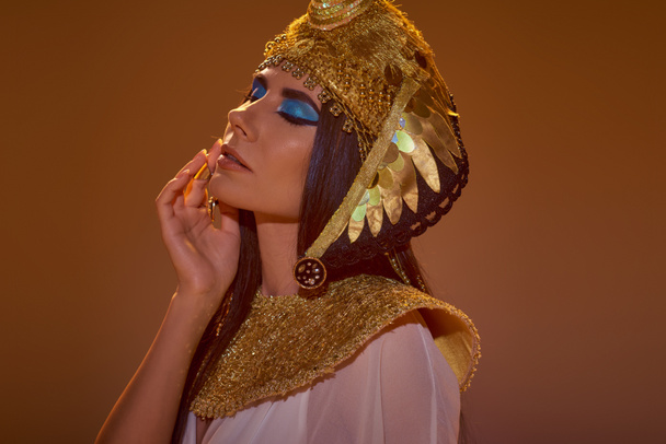 Ritratto di donna con trucco audace e copricapo egiziano guancia toccante isolato su marrone - Foto, immagini