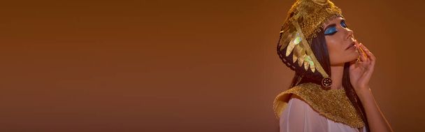 Donna bruna con trucco audace che indossa copricapo egiziano mentre posa isolata su marrone, banner - Foto, immagini