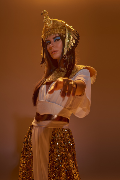 Donna in abbigliamento egiziano punta con il dito alla macchina fotografica e in piedi su sfondo marrone con luce - Foto, immagini