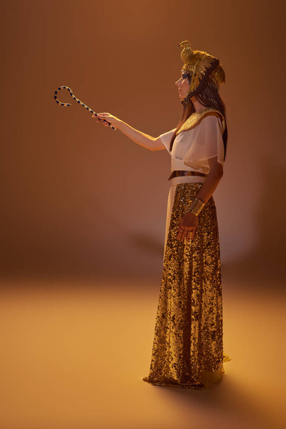 Vista laterale di donna elegante in abbigliamento egiziano tenendo truffatore mentre in piedi su sfondo marrone - Foto, immagini