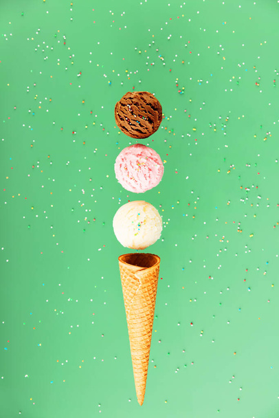 Chocolate, vanilla, strawberry scoops and waffle cone on green background with colorful sprinkles, levitating concept. Spring or summer mood - Фото, изображение