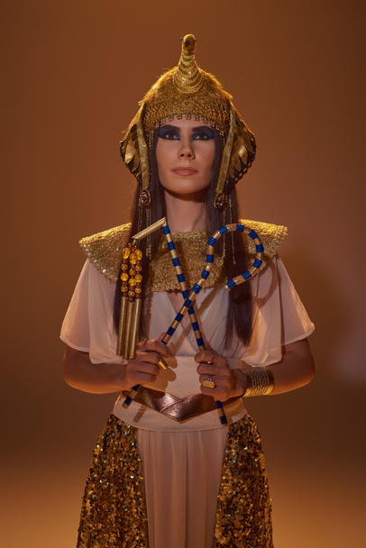 Elegante donna in copricapo egiziano e guardare tenendo truffatore e flagello su sfondo marrone - Foto, immagini