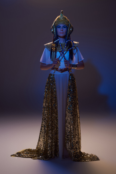 Piena lunghezza di donna elegante in look egiziano tenendo truffatore e flagello su marrone con luce blu - Foto, immagini