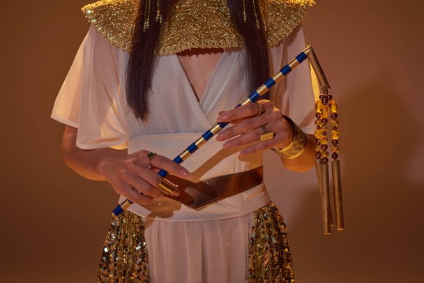 Cropped view of woman in egyptian outfit holding flail while posing on brown background - Photo, Image