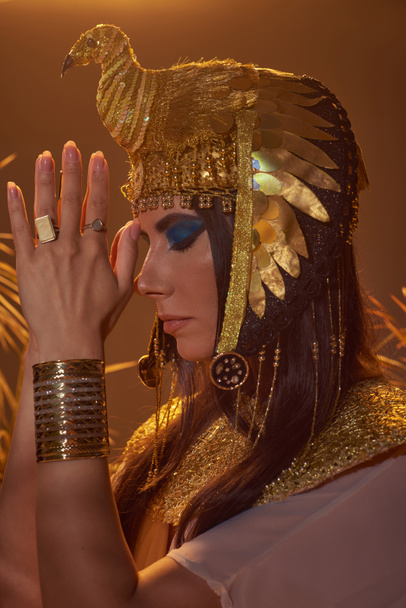 Woman in egyptian costume doing praying hands gesture near blurred plants on brown background - Photo, Image