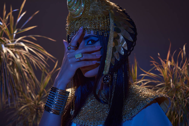 Frau im ägyptischen Look bedeckt Gesicht, während sie in der Nähe von Pflanzen in blauem Licht auf braunem Hintergrund posiert - Foto, Bild