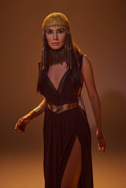 Donna in look egiziano e collana guardando la fotocamera mentre in piedi su sfondo marrone con la luce - Foto, immagini