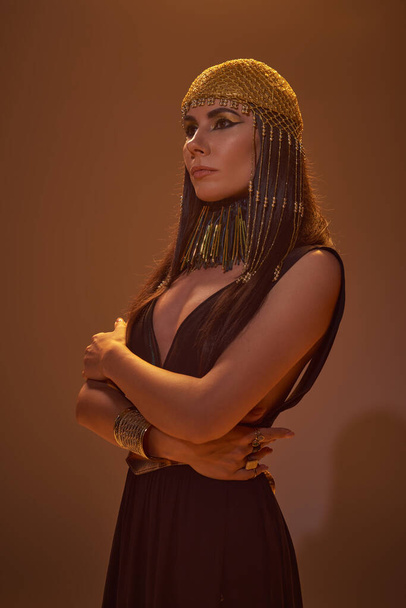 Donna alla moda in look egiziano e copricapo in posa e guardando lontano su sfondo marrone - Foto, immagini