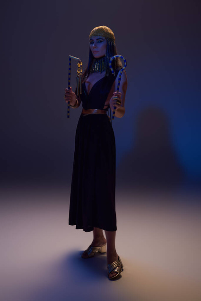 Plná délka ženy v egyptském vzhledu drží bičík a křivák a stojí na hnědé s modrým světlem - Fotografie, Obrázek