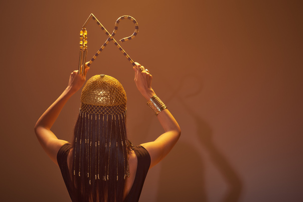 Back view of brunette woman in egyptian headdress holding crook and flail on brown background - Fotografie, Obrázek