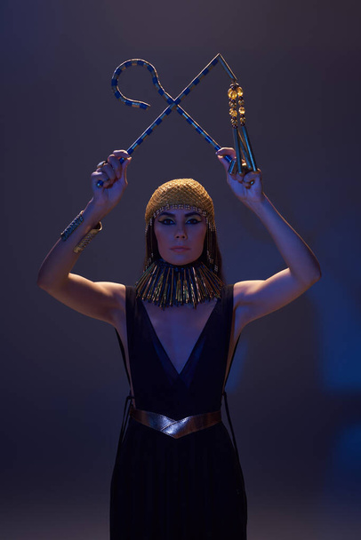 Donna con trucco egiziano e sguardo tenendo truffatore e flagello su sfondo marrone con luce blu - Foto, immagini
