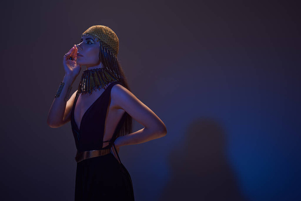 Modelo elegante en atuendo egipcio y tocado posando en luz azul sobre fondo púrpura - Foto, imagen