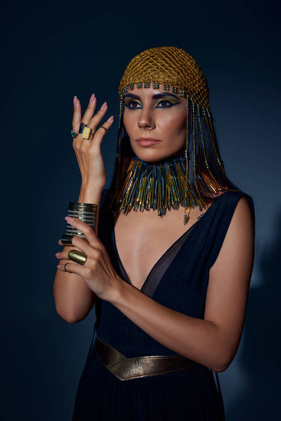 Mujer elegante en mirada egipcia y collar posando mientras está de pie sobre fondo azul - Foto, imagen