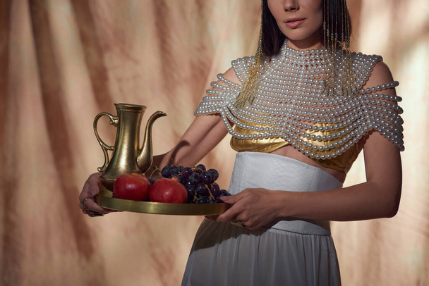 Cropped view of woman in egyptian look holding jug and fruits while posing on abstract background - Photo, Image