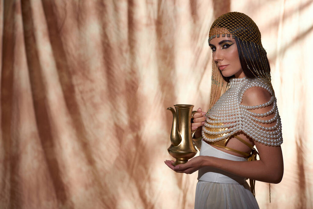 Stylish woman in Egyptian attire and pearl top holding golden jug on abstract background - Фото, изображение