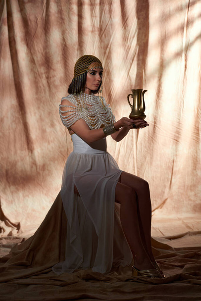 Frau in traditionellem ägyptischen Outfit hält goldenen Krug während sie auf abstraktem Hintergrund sitzt - Foto, Bild
