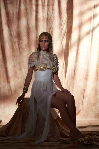 Elegante donna in copricapo egiziano e abbigliamento seduta e in posa su sfondo astratto - Foto, immagini