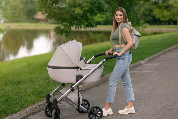 Belle mère avec une poussette marche à travers le parc. Marcher avec un bébé en plein air près du lac. Nouveau-né, famille, enfant, parentalité. - Photo, image