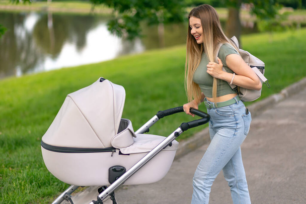 Smiling woman with baby stroller walks through the park near lake. Newborn, family, child, parenthood. - Photo, Image