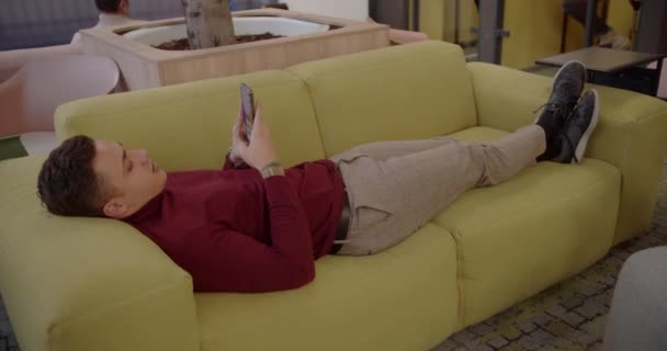 Νεαρός εργάτης αναπαύεται στον αναπαυτικό καναπέ μέσα στο σαλόνι του γραφείου - Πλάνα, βίντεο
