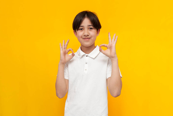 asiático chico de doce años de edad en blanco camiseta muestra gesto de ok en amarillo aislado fondo, coreano niño recomienda y muestra signo de éxito - Foto, Imagen