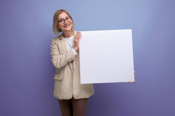 sorrindo mulher madura em casaco demonstrando papel branco mockup no fundo do estúdio com espaço de cópia. - Foto, Imagem