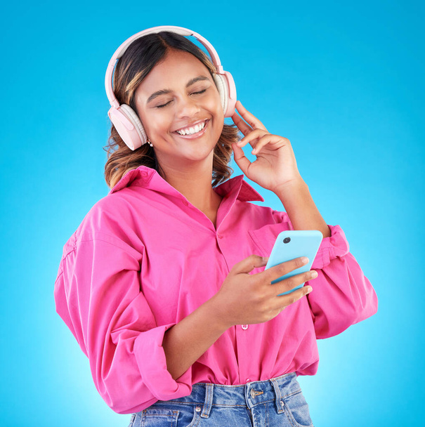 Telefon, hudba a sluchátka se ženou v ateliéru na modrém pozadí, poslouchající rádio. Mobilní, úsměv a šťastná mladá žena streaming audio relaxovat pro wellness nebo svobodu. - Fotografie, Obrázek