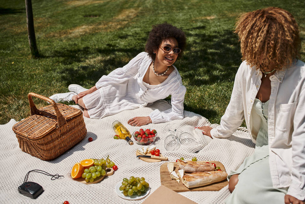 young african american girlfriends talking on blanket near fruits during summer picnic - Fotografie, Obrázek