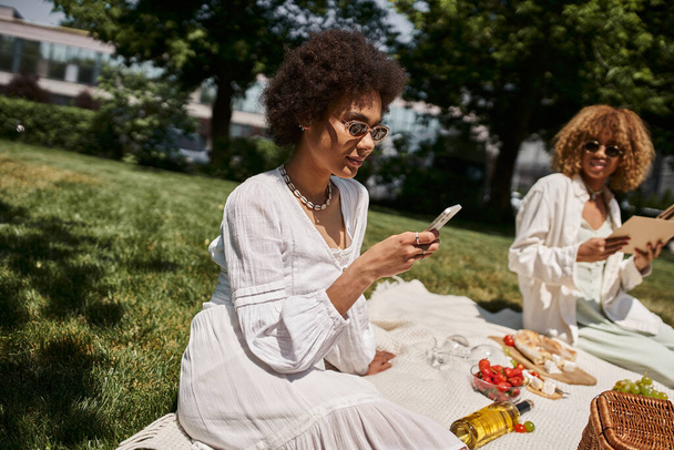 african american woman using smartphone during summer picnic with girlfriend - Photo, Image