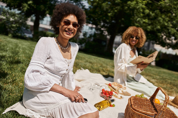 africký americký žena s smartphone pohledu na kameru v blízkosti přítelkyně, letní piknik - Fotografie, Obrázek