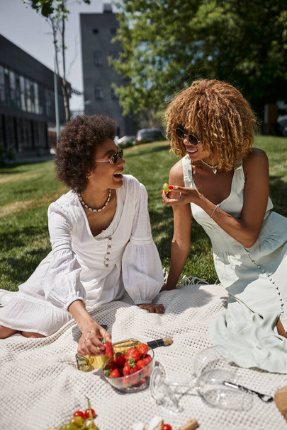 alegres mujeres afroamericanas comiendo frutas y charlando en el picnic de verano en el parque - Foto, Imagen
