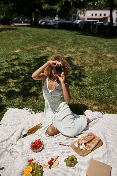 Африканская американка фотографирует на винтажную камеру рядом с вином и клубникой, летний пикник - Фото, изображение