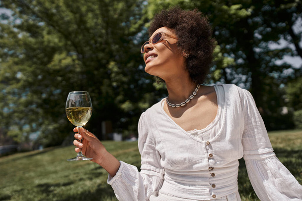 gondtalan és elegáns afro-amerikai nő napszemüvegben kezében pohár bor a nyári parkban - Fotó, kép